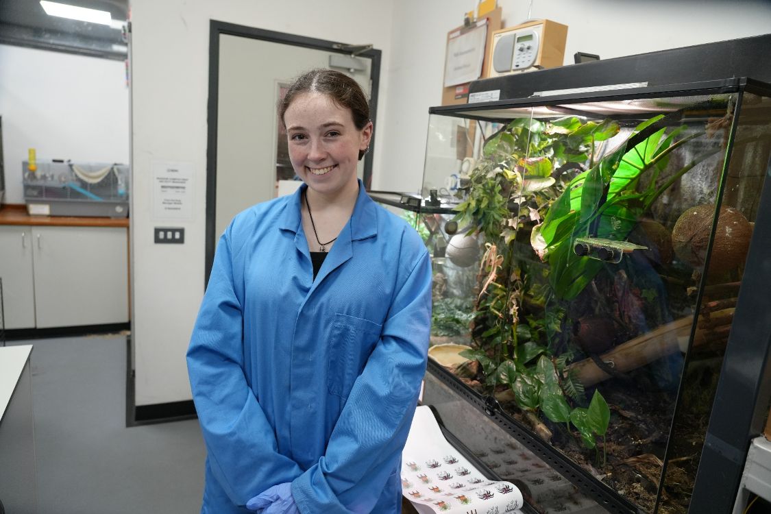 Student Madison Halliday in a blue lab coat standing by a vivarium